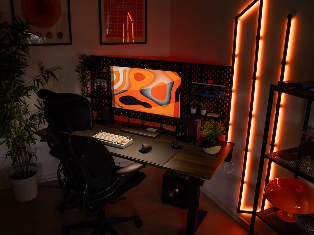Modern gaming setup with a Bifrost standing desk, widescreen monitor, orange LED lighting, and organized accessories.