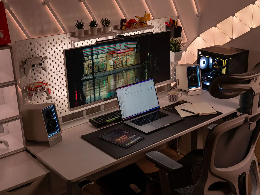 夜晚氛圍的電腦工作桌配置，Dezctop Bifrost白色升降桌搭配大型螢幕、筆電與平板，後方洞洞板整合手把架與收納盒，兩側擺放藍光音響與電腦主機，搭配人體工學椅與間接燈光，營造沉穩科技感的工作空間。