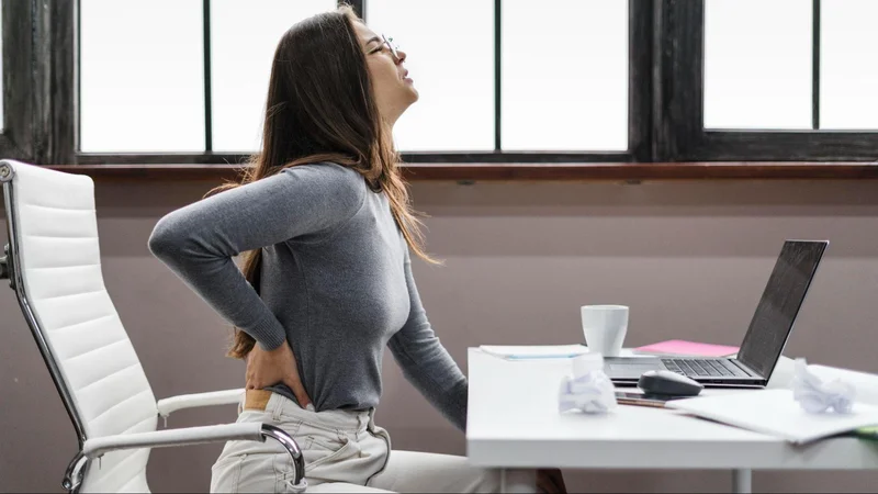 Captured in an office environment, the photograph showcases a woman seated at a desk, her back turned away from the camera. This image highlights the significance of workplace wellness and the need to adopt healthy back habits.